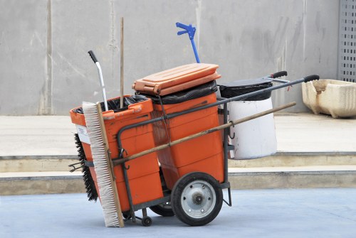 A wheeled orange plastic rubbish bin with a black lid, positioned on a light grey pavement surface in front of a concrete wall. The bin is filled with various cleaning tools and equipment, including a long-handled broom with black bristles and silver handle resting against its side, a blue-handled tool or event marker, and a smaller black object on top. Nearby, a pair of brown flat objects, possibly cushions or floor mats, are propped against the lid, and a white container is attached to the side of the bin. In the background, there is a large beige plastic container lying on its side and a metallic or perforated grey panel. The scene suggests a setting for waste collection or cleaning activities, consistent with services offered by Commercial Waste Gipsy Hill, in an urban or commercial area of Gipsy Hill with clean, well-maintained surroundings.