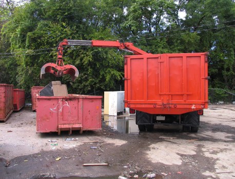 The image depicts a waste collection operation outdoors, featuring a red industrial waste grab attachment mounted on a hydraulic arm, which is actively lifting or emptying waste into a large, open-top red skip or container. The skip is positioned on a paved or gravel surface, with visible debris and litter scattered around the area. In the background, there is a row of dense green trees and greenery, indicating a location possibly near a park or a roadside setting. To the left, part of another similar red waste bin or container is partially visible. The scene suggests an organized rubbish removal activity, typical of commercial waste services such as those provided by Commercial Waste Gipsy Hill, serving areas around Gipsy Hill and the surrounding postcode regions. The natural daylight and shadows imply the photo was taken during daytime, emphasizing the industrial equipment and waste materials involved in the rubbish collection process.