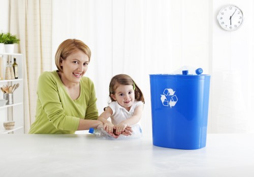 A woman with short blonde hair wearing a light green top is sitting at a white table alongside a young girl with dark hair styled in pigtails, dressed in a white top. They are both smiling and appear to be engaging in a rubbish disposal activity, with the woman holding a clear plastic bottle. On the table, there is a large, bright blue recycling bin featuring a white recycling symbol, positioned to the right of the pair. The background shows a bright interior space with white walls, a wall-mounted clock, and a shelf with decorative items, suggesting a domestic environment. The scene depicts waste management practices, possibly related to household rubbish collection services in Gipsy Hill, with a focus on recycling and waste separation.