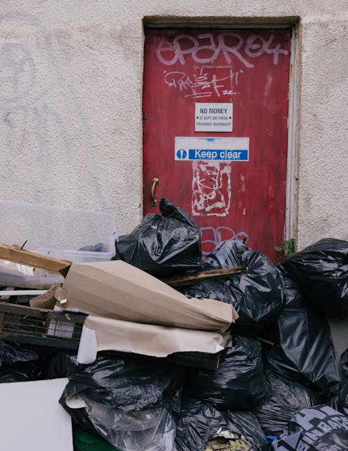 A pile of black rubbish bags, some partially torn open, are stacked in front of a red metal door on a concrete wall. The bags contain discarded waste and are surrounded by various items including card