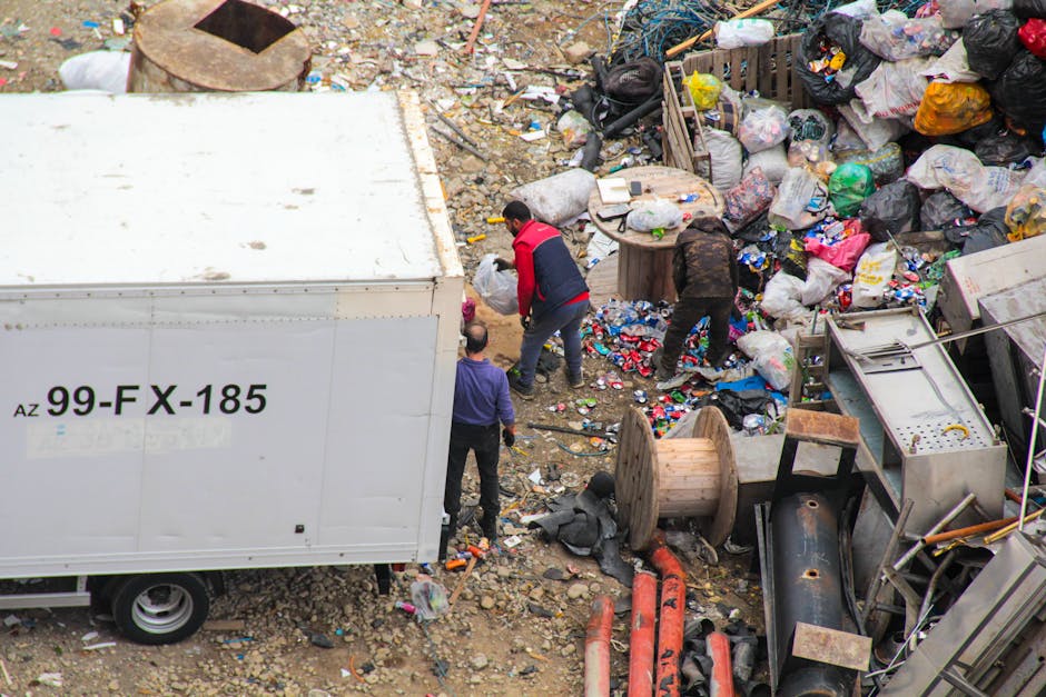 This image depicts a large accumulation of mixed waste and rubbish overflowing from several black and red wheelie bins placed on a paved urban street. The waste includes crumpled paper, cardboard boxe