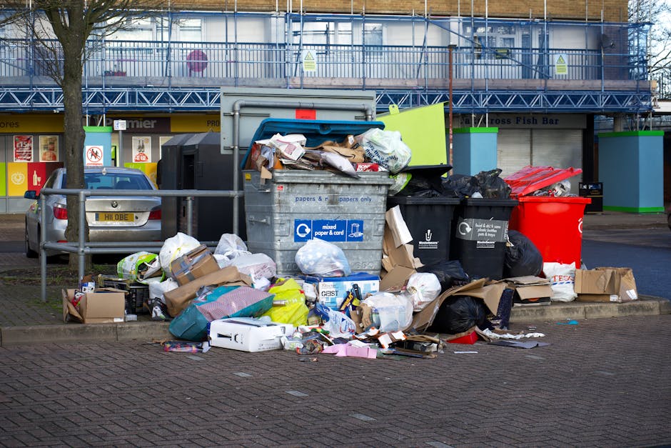 A pile of mixed waste materials and rubbish bags sits on a gravel surface outdoors, positioned near a low stone wall. The debris includes black plastic garbage bags, a yellow plastic storage container